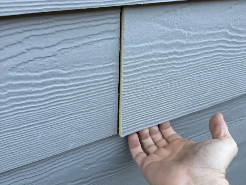 An inspector's hand lifting a loose piece of lap siding away from the wall of a new home